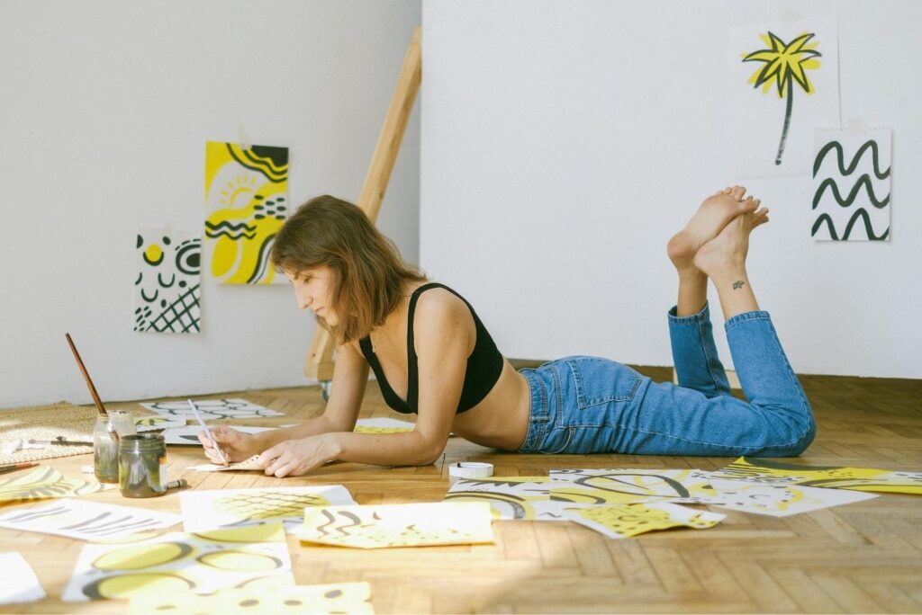 A woman lying on the floor on her stomach, working on abstract, white, black, and yellow paintings.