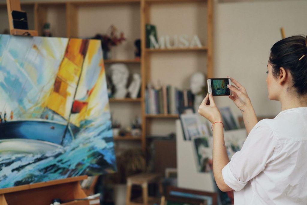 An artist in her studio using her cellphone to take a photograph of a finished painting.
