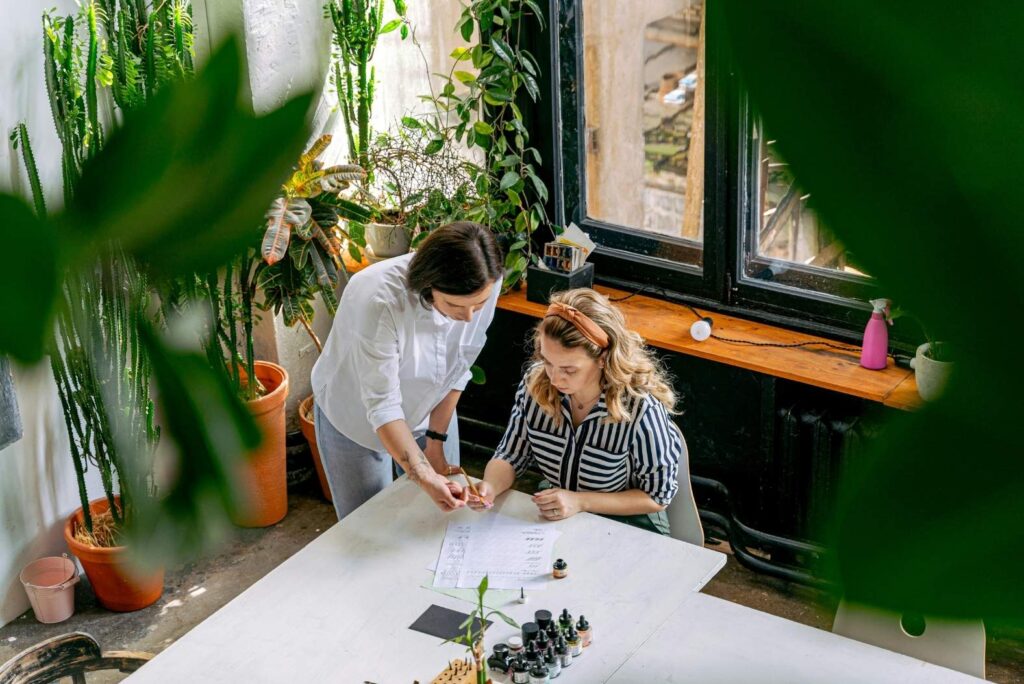 Two women sitting in a sunlight room full of plants. They're working together on an art project.