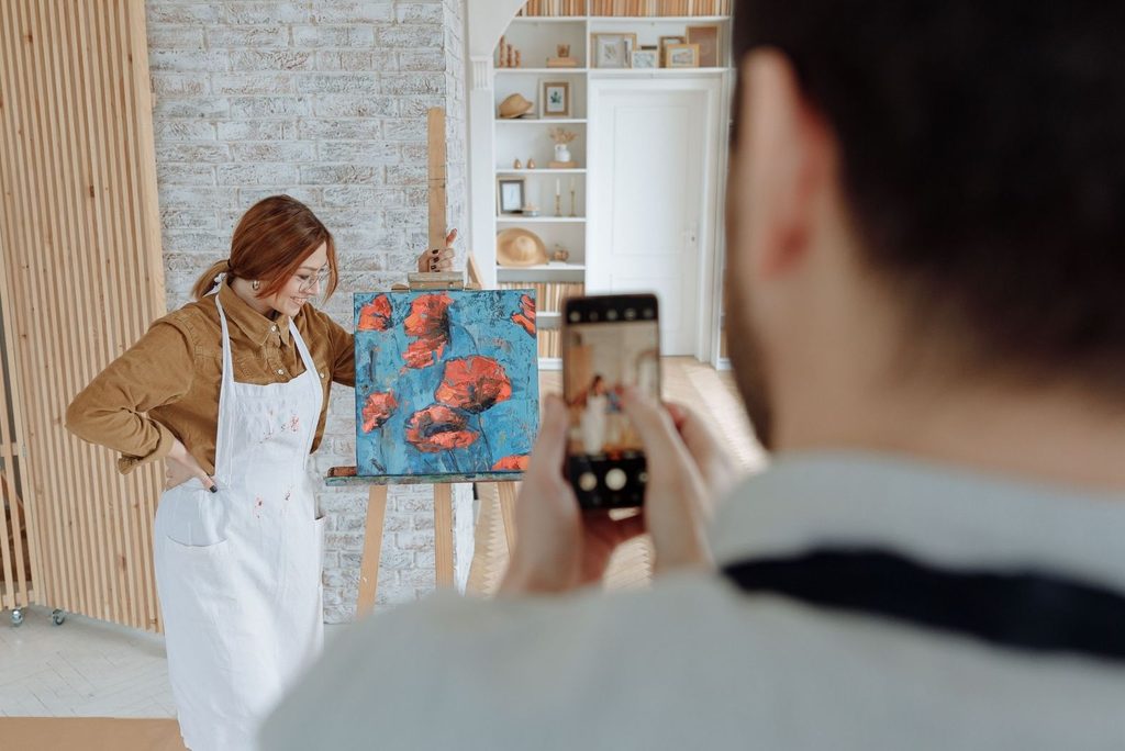 An artist in her studio posing next to a painting she just completed. A man is taking a photo of her with his phone.