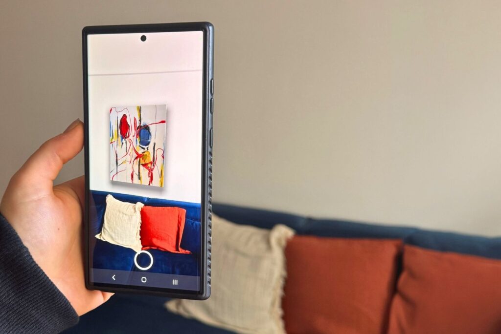 A hand holding a phone in a living room. On the phone, the person is trying out a piece of abstract art with Augmented Reality. They can see what the painting would look like in their own room and decide if they want to buy.
