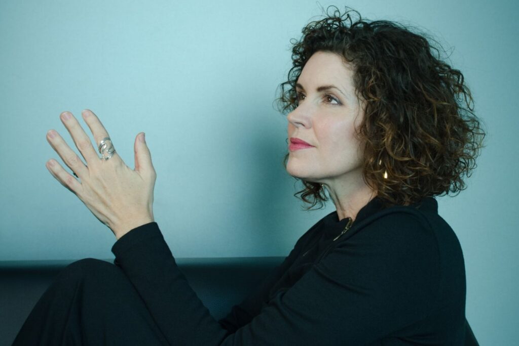 Artist headshot of Crista Cloutier, founder of The Working Artist. Sheâs sitting in profile against a solid, light blue background, looking wistfully above. Thereâs a calm, reflective feeling to the photograph.