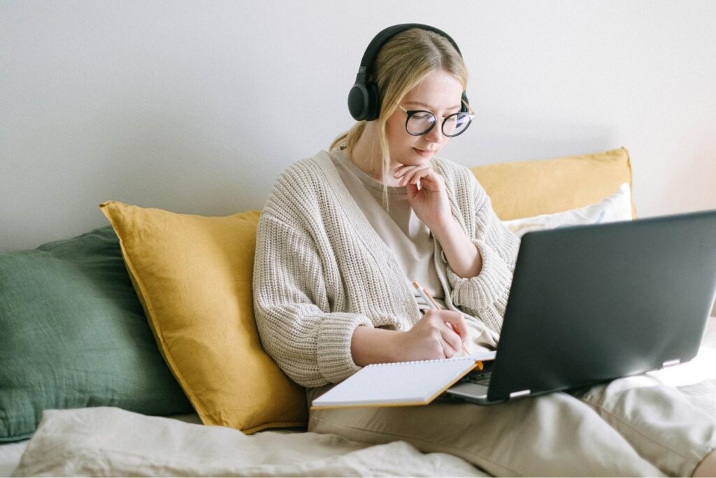 Woman wearing headphones and glasses sits on a couch with yellow and green pillows, writing in a notebook while looking at a laptop screen.