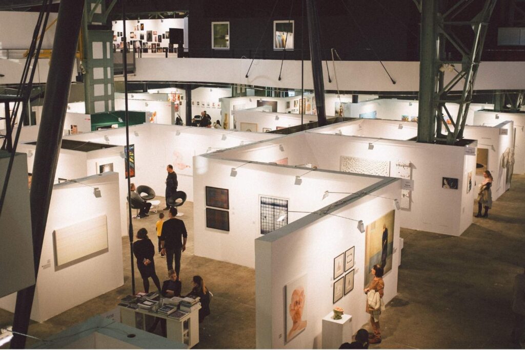 Aerial view of an art fair with visitors walking between white booths hung with artworks.