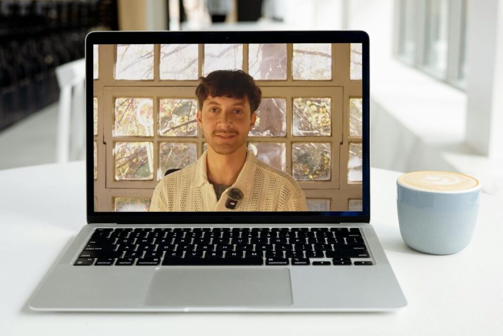 Laptop on a desk showing a video call with a smiling man, next to a cup of coffee.
